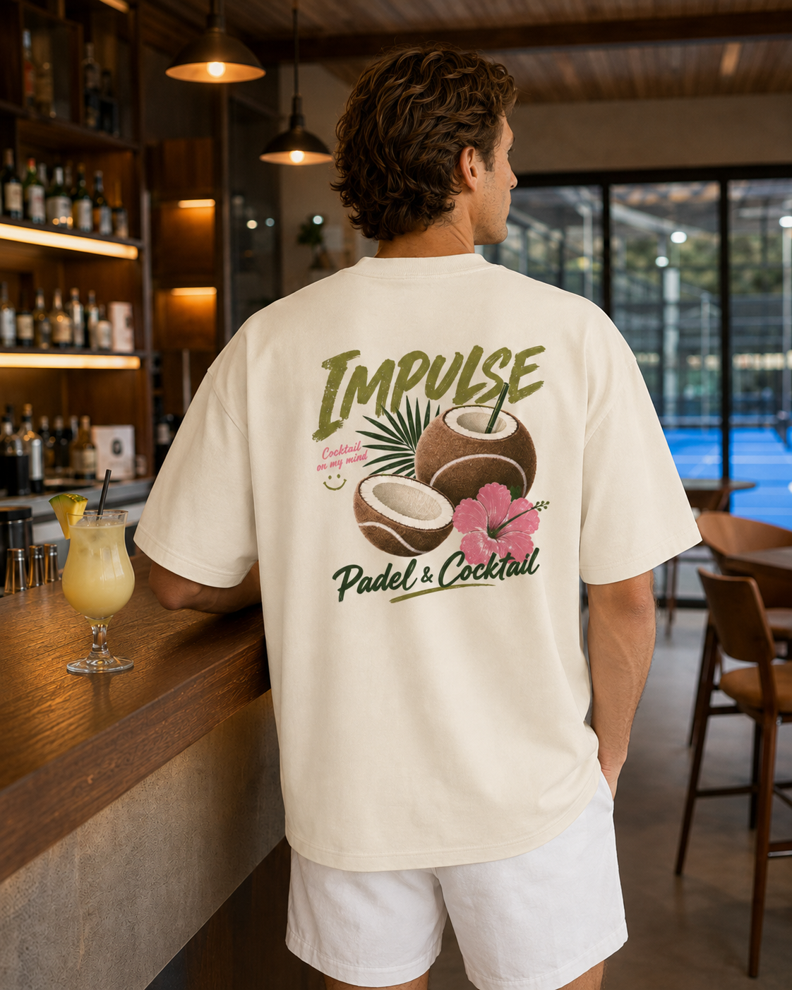 T-shirt homme oversize Padel & Cocktail ivoire porté de dos au bar d’un club de padel