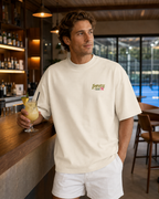 T-shirt homme oversize Padel & Cocktail ivoire porté de face au bar d’un club de padel
